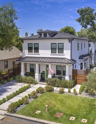 A two-story white house with black accents, an American flag, and a well-manicured lawn, flanked by other houses and trees in a residential neighborhood.
