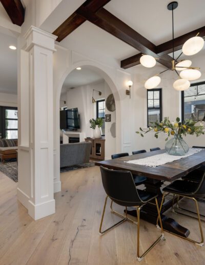 Modern open-concept dining and living area with a wooden table, black chairs, light wood flooring, archway, and a contemporary chandelier. TV and seating in the background.