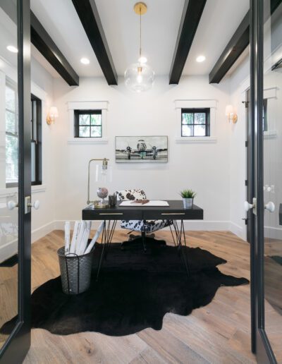A minimalist home office features a black desk, chair, and cowhide rug, with decorative items on the desk. Wooden flooring and black ceiling beams complement the neutral wall tones.
