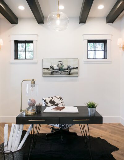 A modern home office with a desk, lamp, globe, and plant on a black rug. Artwork of an airplane is on the wall. Glass doors and windows bring in natural light.