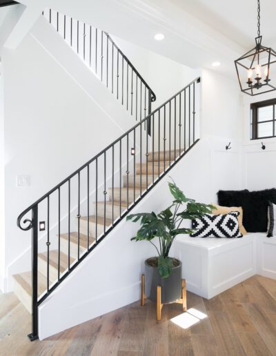 A modern entryway features a staircase with black railing, wooden floors, a built-in bench with black and white cushions, and a potted plant. Pendant lights hang from the ceiling.