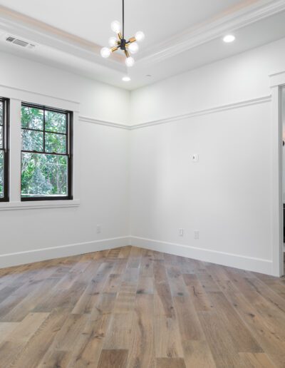An empty room with wooden floors, white walls, two windows, and a modern light fixture. A doorway leads to a bathroom with visible sinks and a mirror.