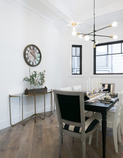 A modern dining room with a black and white table set for six, a round wall clock, a side table with plants, and a contemporary chandelier. Wooden flooring and large windows are visible.