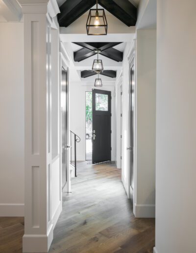 A hallway with wooden flooring leads to a black front door, featuring white walls, modern light fixtures, and ceiling beams.