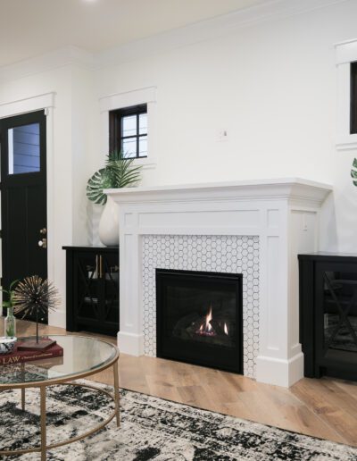 A living room with a modern fireplace, black and white decor, round glass coffee table, and potted plants on mantel.