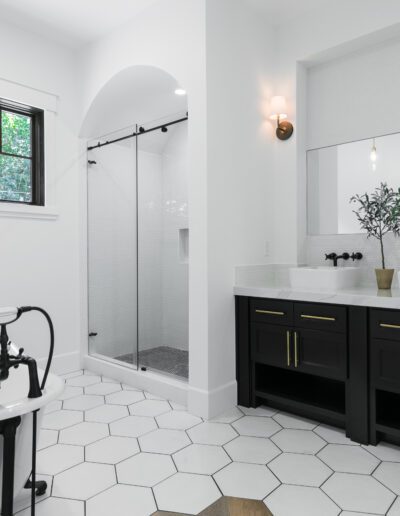 Modern bathroom with a freestanding bathtub, double sink vanity, large mirror, and glass-enclosed shower. White walls and hexagonal floor tiles. A small plant decorates the sink area.