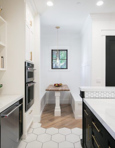 Modern kitchen with white hexagonal floor tiles, black lower cabinets, white upper cabinets, a farmhouse sink, and a small dining nook with a wooden table and bench under a window.