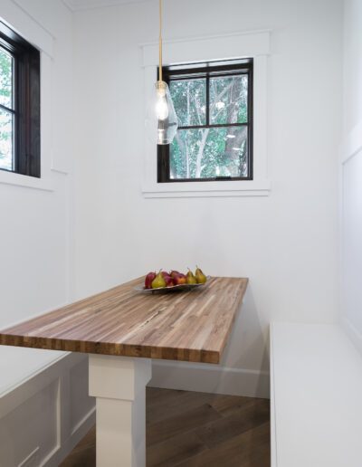 A minimalist dining nook with a wooden table, white benches, and a hanging light. A bowl of fruit is on the table. White walls and two windows complete the space.