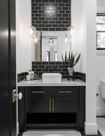 A modern bathroom with black subway tiles, a white sink on a black vanity, round mirrors, pendant lights, and a small plant. Black and white floor tiles and a toilet are visible.