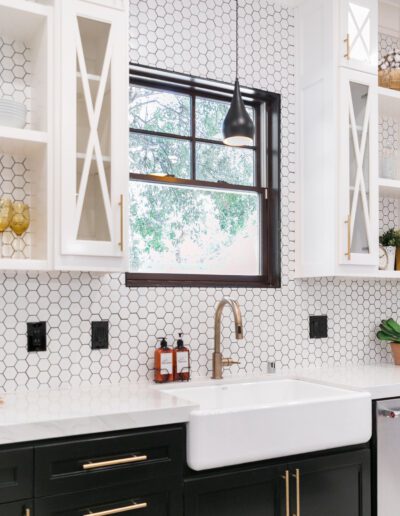 Bright kitchen with hexagonal tiled backsplash, white cabinets, black lower cabinets, farmhouse sink, and a pendant light over the countertop. A window shows trees outside.