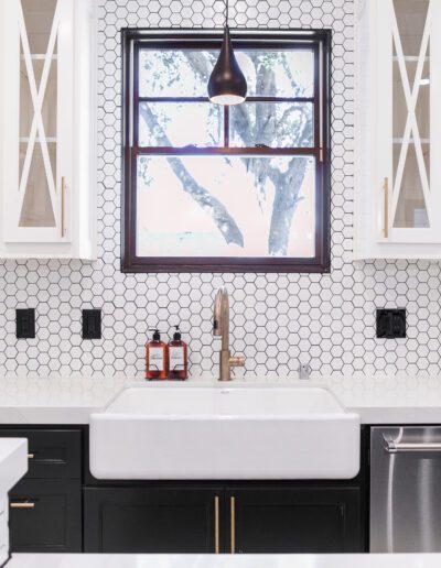 A modern kitchen with white hexagonal tile backsplash, white farmhouse sink, dark cabinets, open shelves with decor, and a window above the sink.