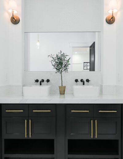 A modern bathroom features a double vanity with black cabinets, white sinks, brass fixtures, and a central plant in a pot, flanked by two wall sconces.