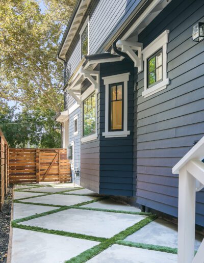 Side view of a blue house with white trim, featuring a paved walkway with grass strips, wooden fence, and stairs leading to a door.