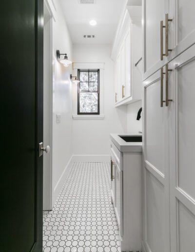 Narrow laundry room with white cabinets, hexagonal floor tiles, a window, sink, and black door.