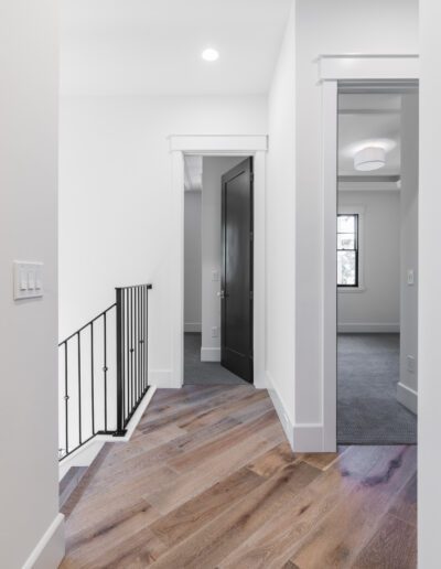 A hallway with wooden floors, a black door, and a staircase with a black railing, leading to a carpeted room.
