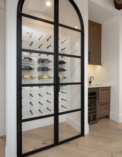 A modern kitchen with a large black-framed glass door wine rack, adjacent to a bathroom and a dining area with bar stools and pendant lights.