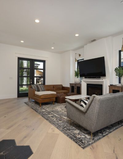 A modern living room with a sofa, armchair, and ottoman. A large TV is mounted above a fireplace. The room has a rug, wooden floor, and various wall mirrors and decor.