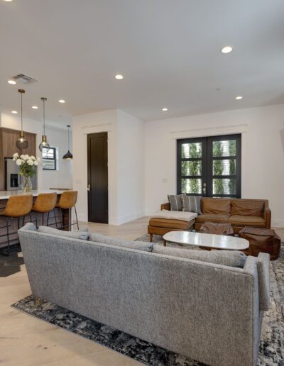 Modern open-concept living area with a gray sofa, leather chairs, and a TV on the wall. The kitchen features wood cabinets, an island with stools, and pendant lighting.