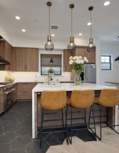 Modern kitchen with wooden cabinets, a central island with three brown barstools, pendant lights, and a black hexagonal tile floor. White countertops and stainless steel appliances are featured.