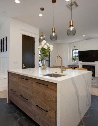 Modern kitchen with a marble island, pendant lights, and wood accents. Open living area with a TV, armchair, and decorative items on walls and shelves. Light wood flooring.