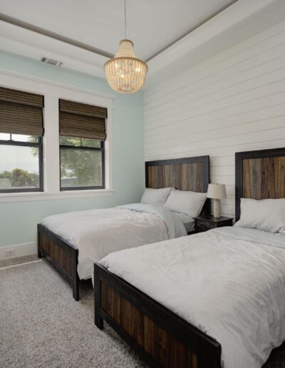 Bedroom with two wooden twin beds, white bedding, a small nightstand, and a hanging light fixture. Walls are light blue with white shiplap panel and two windows with brown blinds.