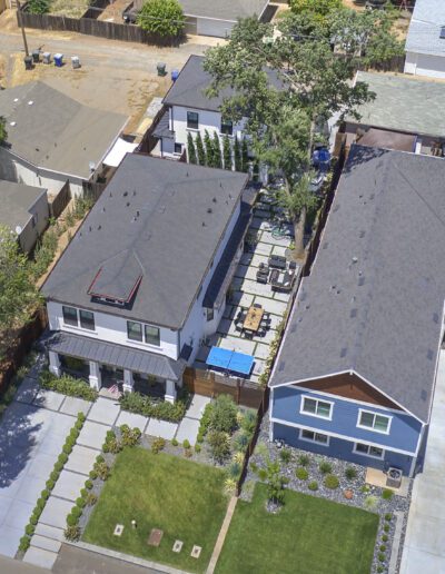Aerial view of residential neighborhood with two adjacent houses, neatly manicured lawns, and a central shared patio area. Several cars are parked in driveways.