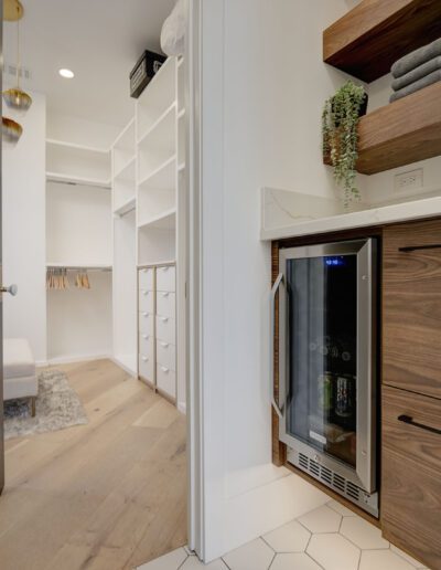 View of a modern walk-in closet with wooden shelves and a mini fridge. The closet features white walls, gold light fixtures, and ample storage space with drawers and hanging rods.