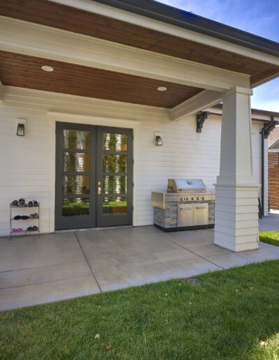 Outdoor patio area with a covered porch, glass double doors, a built-in grill station, and neatly arranged shoes on a rack. Surrounded by fenced yard and greenery.
