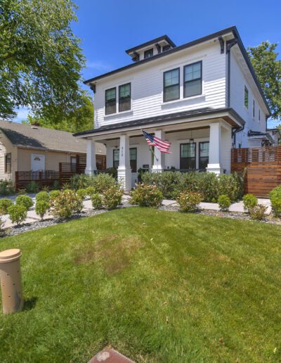 A two-story white house with an American flag on the porch, surrounded by a manicured lawn and shrubs, under a clear blue sky.