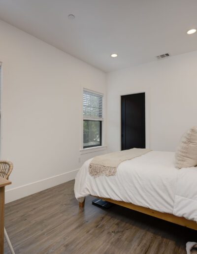 Minimalist bedroom with a single bed, white bedding, a wooden desk with flowers, a wicker chair, and two windows with blinds. Walls and ceiling are white; floor is wood.