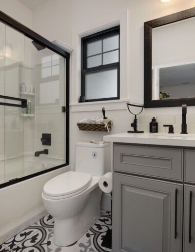 A bathroom with a glass-enclosed bathtub, gray vanity, white countertop, large mirror, toilet, and patterned tile floor.
