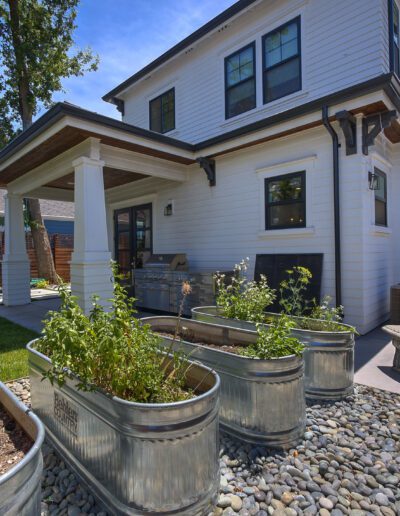 Backyard view of a two-story house with a covered patio, metal planters filled with plants, a stone picnic table, and surrounding greenery.