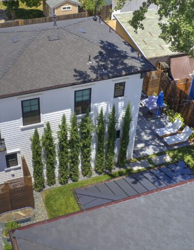 Aerial view of a two-story house with a gray roof, white siding, and tall trees lining one side. Adjacent are a patio, outdoor furniture, and a tree providing shade.
