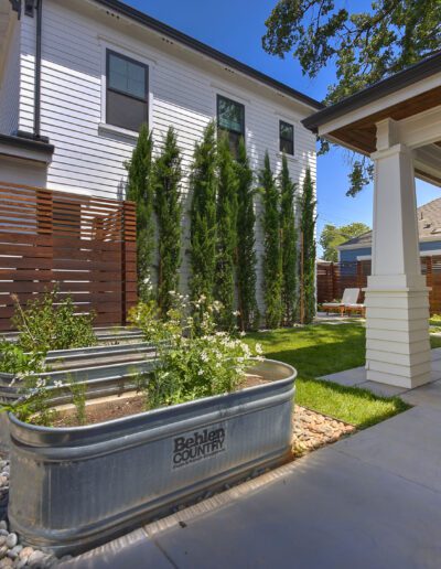 Modern backyard with a white house, metal planters, a lawn, and a covered patio with seating. Surrounded by a wooden fence and landscaped with rocks and vertical greenery.