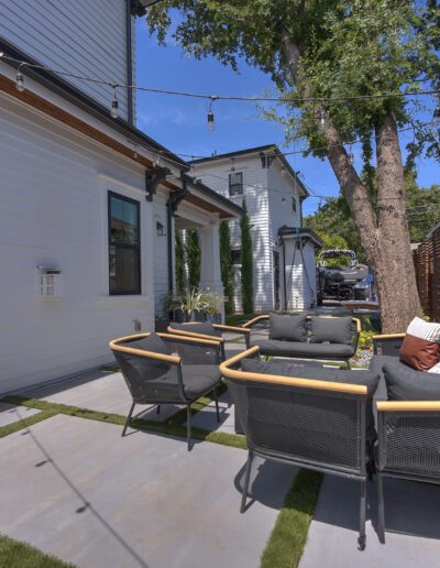 Outdoor patio area with modern patio furniture, string lights overhead, and a wooden privacy fence. A tree and neighboring house are visible in the background.
