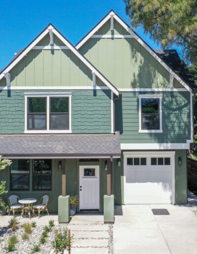 A green two-story house with a gray roof, garage, and small front patio with table and chairs, surrounded by trees and a gravel driveway.