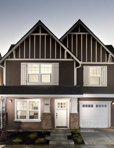 A two-story house with a gabled roof, brown siding, and white trim. It has a single garage, front porch, and illuminated windows. The driveway and garden are visible in the foreground.