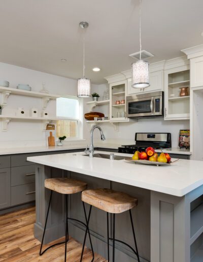 Modern kitchen with gray and white cabinetry, an island with a white countertop, two wooden stools, stainless steel appliances, open shelves, and pendant lighting above the island.