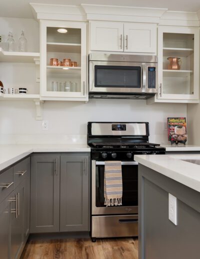 Modern kitchen with white and gray cabinets, a gas stove with an overhead microwave, a sink island, and wooden flooring. Shelves and countertops display decor and utensils.