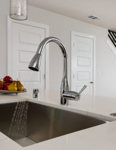 A modern kitchen with a sleek faucet spraying water into a stainless steel sink. A bowl of apples and pears sits on the white countertop. A staircase and door are visible in the background.