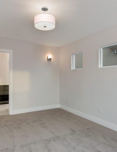 Empty bedroom with gray carpet, white walls, a ceiling light, and a door leading to a bathroom with a vanity and mirror. Three small windows are on one wall.
