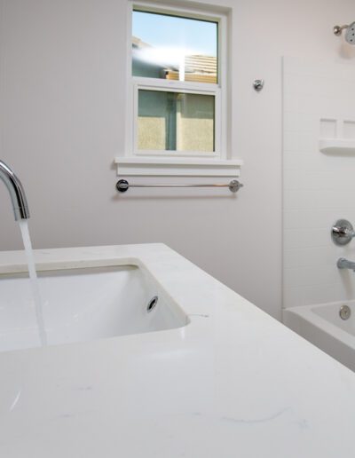A modern bathroom with a white countertop, sink with running water, a bathtub, shower, and a window.