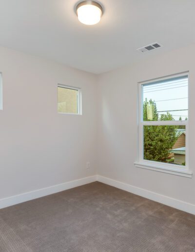 Empty room with beige carpet, white walls, two small square windows, and one rectangular window. Ceiling light fixture and wall power outlets visible.