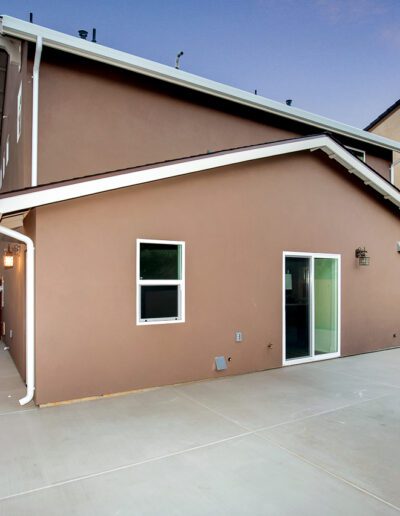Backyard view of a modern two-story house with beige exterior, small windows, sliding glass door, air conditioning unit, and fenced yard. The sky shows a gradient of pink and blue at dusk.