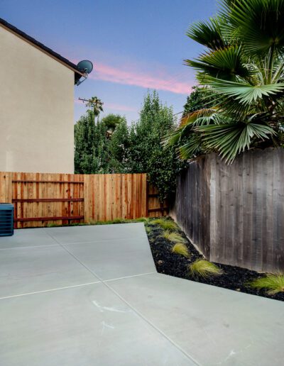 A backyard with a concrete patio, a wooden fence, and a small garden area featuring palm trees and shrubs. An air conditioning unit is installed near the house.