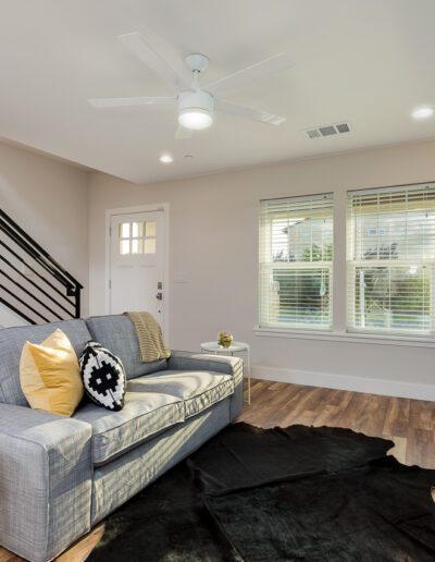 Modern living room with a gray sofa, decorative pillows, a globe on an end table, a black rug, staircase, ceiling fan, windows, and a white fireplace.